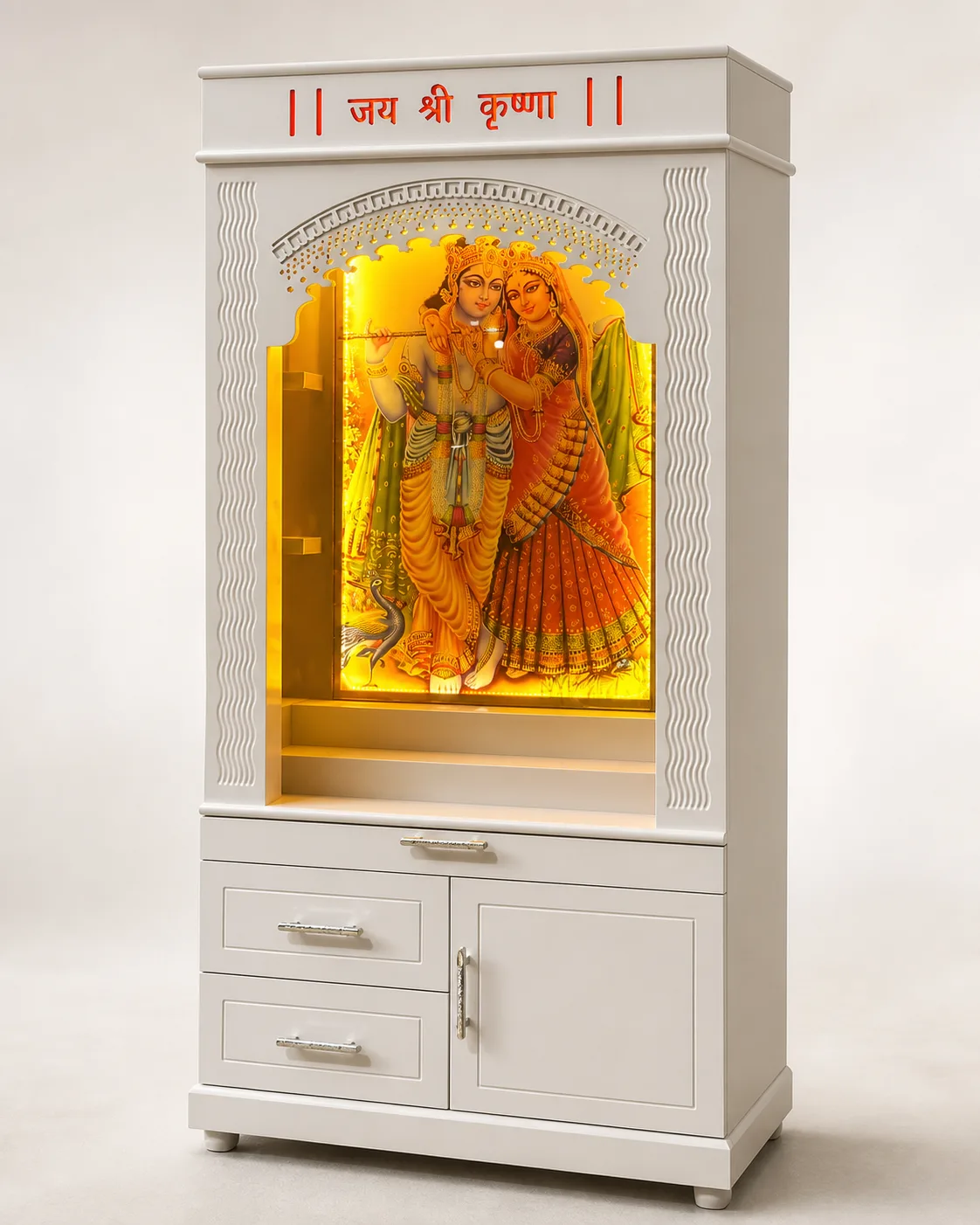 White mandir unit with an amber-lit niche holding a Radha-Krishna print panel and fluted side pillars.